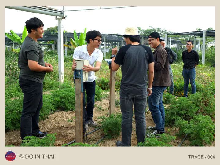 A group talks around a device among planted rows beneath a roof frame.