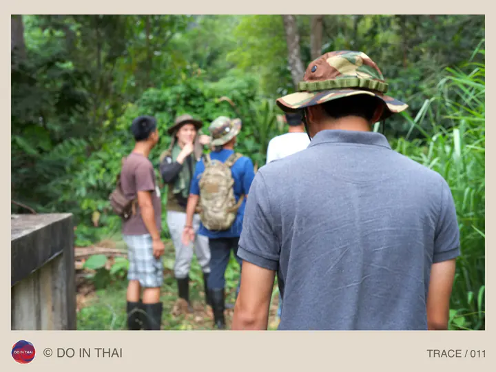 A person in a patterned hat follows a group along a narrow wooded path.