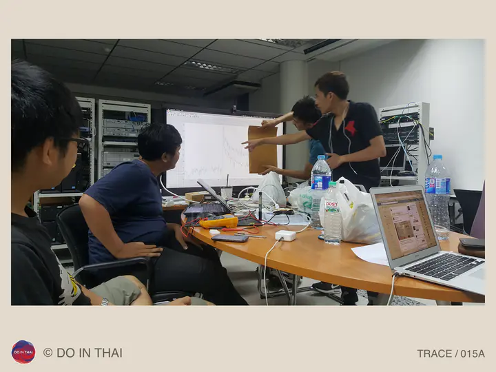 Team members point towards a projected graph beside a table crowded with laptops, wires, and electronic equipment.