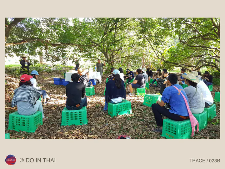 A group sits on green crates under orchard trees, facing speakers and a board at the front.