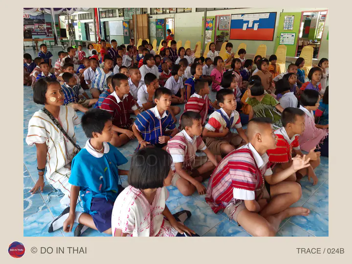 Children in colourful striped shirts sit together on a blue tiled floor in a school hall; several are clapping.