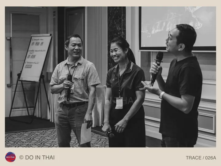 Black-and-white photograph of three people holding microphones and smiling beside a flip chart and projection screen.