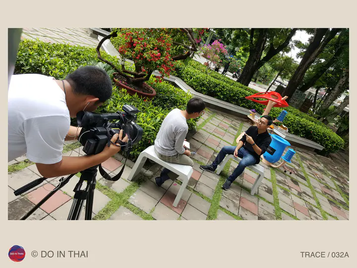 A camera operator films two people talking on outdoor benches beside a colourful tree-shaped weather instrument.