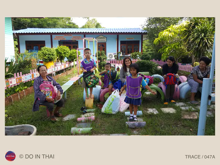 Children with colourful backpacks and adults pose in a school garden, beside painted tyre planters and a blue building.